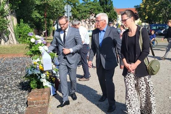 Drei Menschen tragen einen Kranz zur Gedenkstätte.