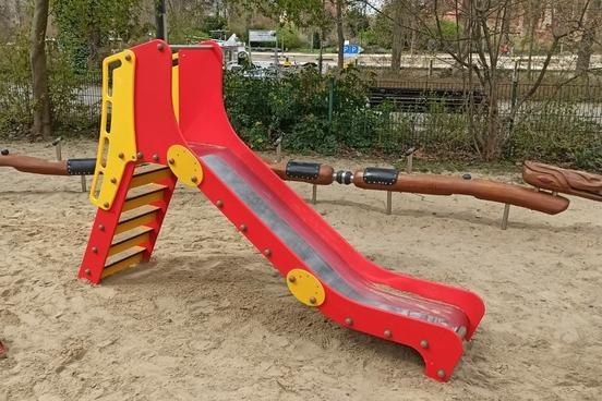 rot-gelbe Kleinkindrutsche auf dem Sandboden des Spielplatzes, im Hintergrund das Schlangenspielgerät