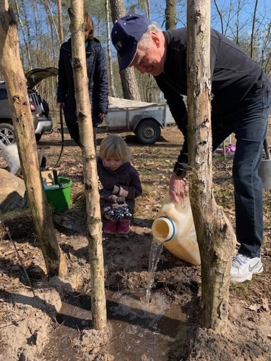 ein Mann schüttet Wasser aus einem Wasserkanister an den Stamm der eingepflanzten Birke, ein kleines Kind und eine Frau schauen zu