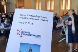 Flyer der Veranstaltung im Vordergrund, dahinter eine Sprecherin am Pult und das Publikum im Rolandsaal