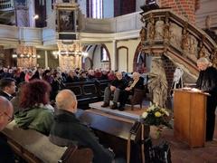 zwei ältere Personen sitzen auf einer kleinen Couch in der St. Gotthardtkiche, ein Mann steht am Rednerpult daneben