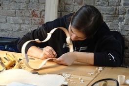 Eine Frau fertigt eine Geige, der Rohkörper der Geige liegt vor ihr auf einem Tisch.