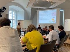 Mehrere Personen sitzen an einem Tisch und schauen auf eine PowerPoint Präsentation während eine Frau dazu spricht.