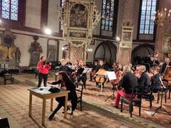 Musiker mit Streichinstrumenten sitzen auf der Bühne, die Dirigentin sitzt vor einer Schreibmaschine