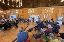 Viele Gäste sitzen in einer Art Stuhlkreis um das Rednerpult herum und beobachten eine Frau die davor steht und redet.