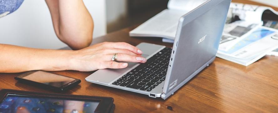 Laptop, Handy und Tablet liegen auf einem Tisch, Hände einer Person sind zu sehen