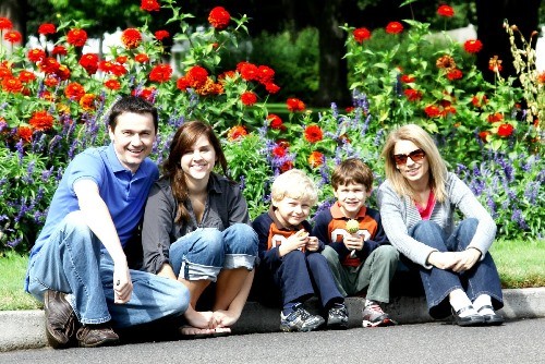 Familie sitzt auf einer Bordsteinkante und lächelt, im Hintergrund blühende Blumen
